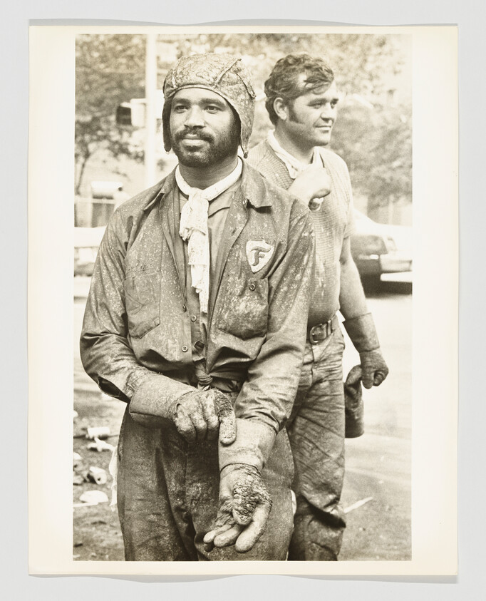A worker wearing a protective cap shows heavily soiled, callused hands while another worker stands behind him.
