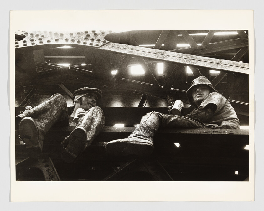 Two construction workers sit on a steel beam high above the ground under a metal framework.