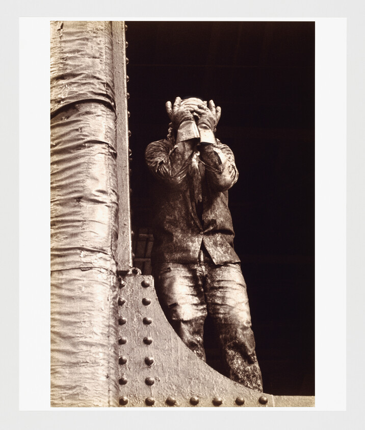 A heavily dust-coated worker stands by a riveted steel beam, covering their face with both hands.