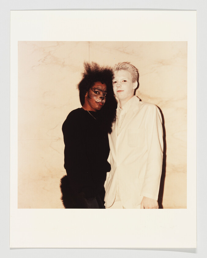 Two people stand close together, one wearing a dark furry costume and face makeup, the other in a white suit.