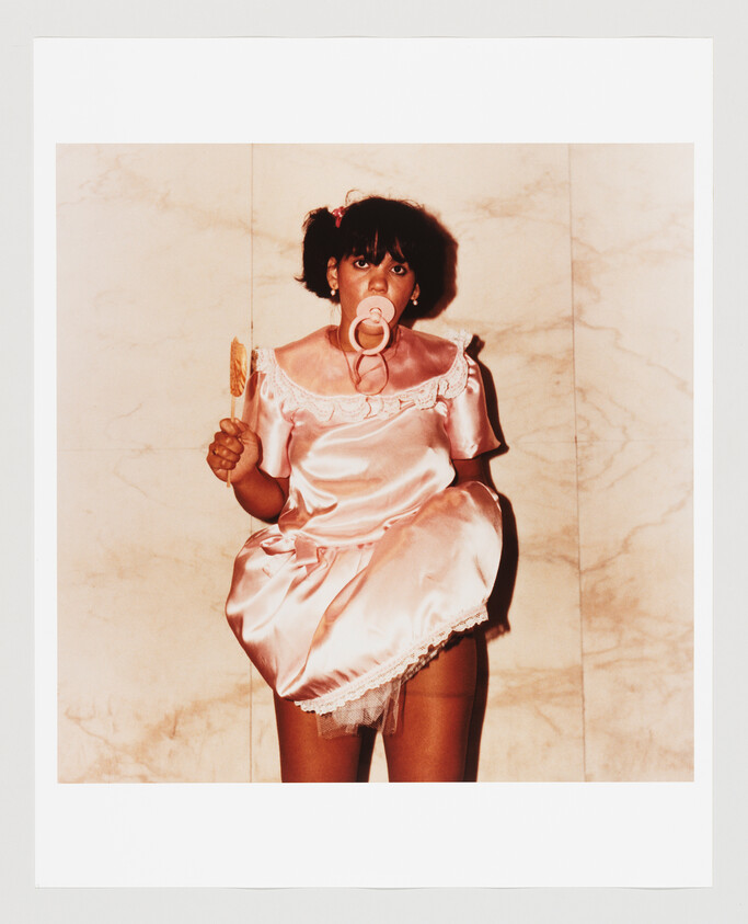 Person dressed in a pink baby dress with a pacifier in their mouth holding a lollipop.