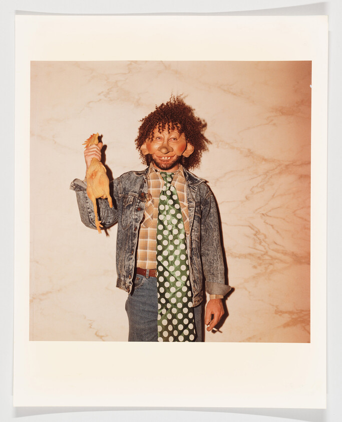 Person wearing a quirky mask and denim jacket holds a rubber chicken while standing against a wall.