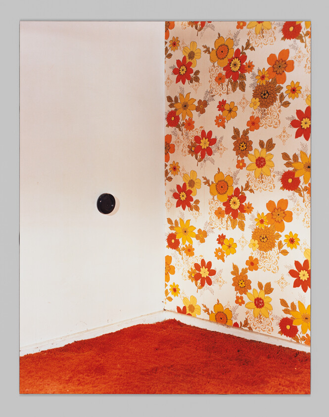A room corner with bright orange floral wallpaper, white wall with a round vent, and orange carpet.