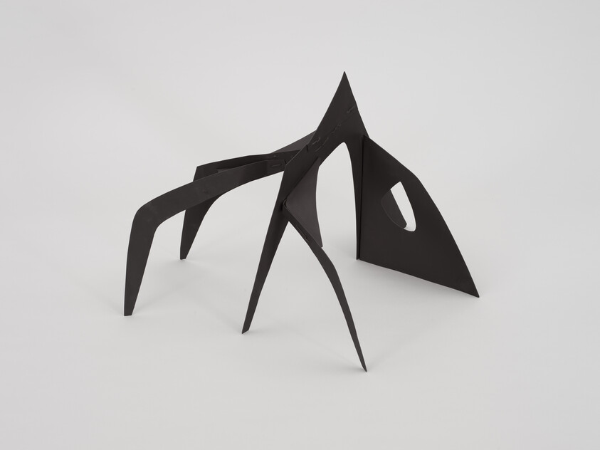 Black abstract metal sculpture with four pointed legs and a cut-out hole on one side, set on white background.