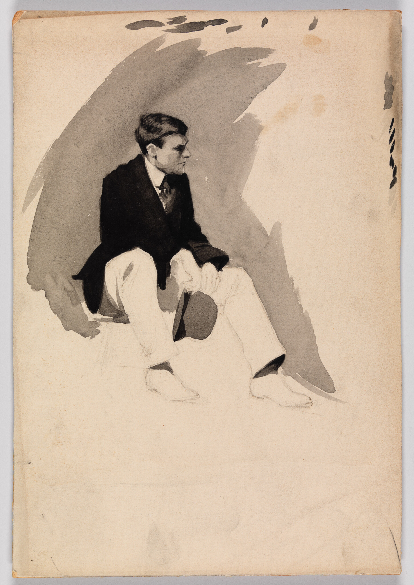 A young man in a dark jacket and light trousers sits holding a hat and looking right.