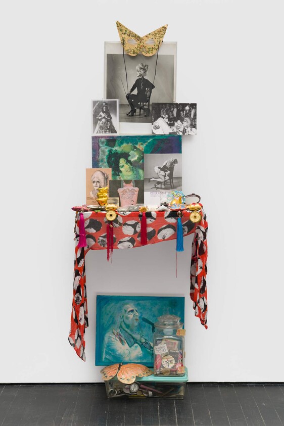 Small wall shrine with photographs, a gold mask, colorful cloth, and assorted trinkets on a shelf.