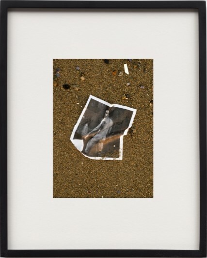 A torn black-and-white photograph of a person lies on wet sand among small pebbles.