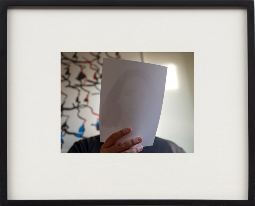 Person holds a sheet of paper with a faint printed face obscuring their own face.