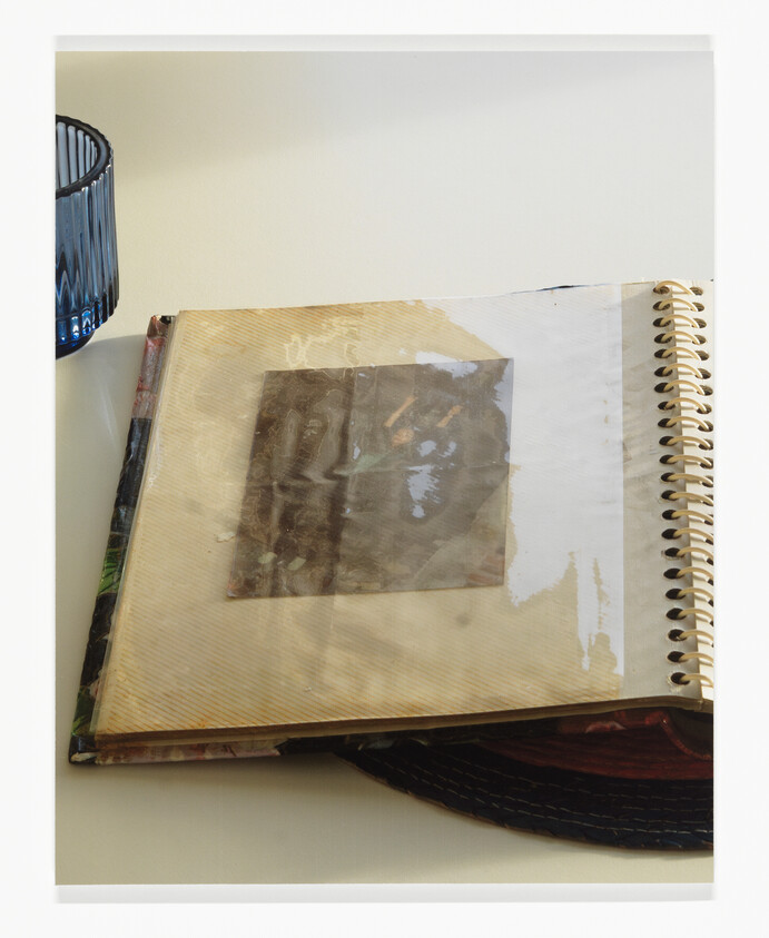 A spiral-bound scrapbook lies open on a table showing a taped photograph, with a glass nearby.