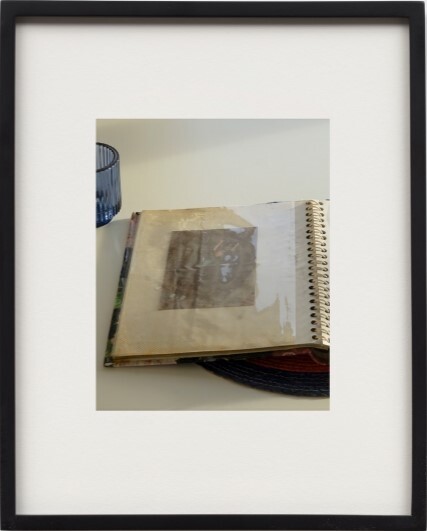 A spiral-bound scrapbook lies open on a table showing a taped photograph, with a glass nearby.