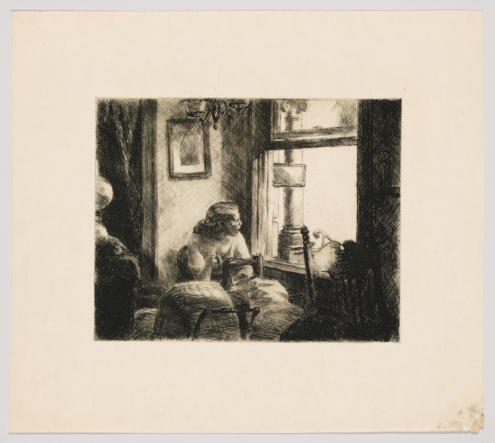 A woman sits at a sewing machine by a window, gazing outside.