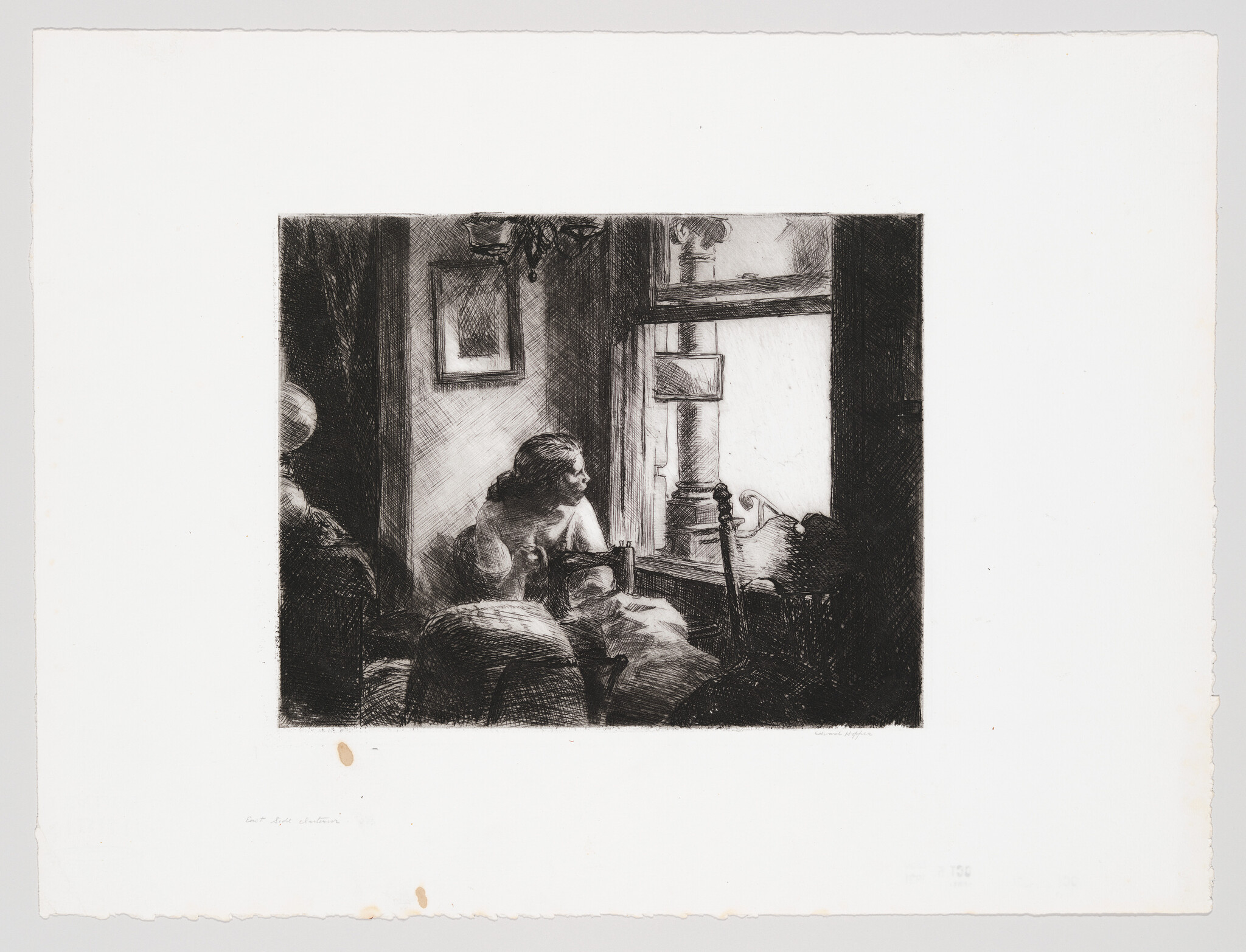 A woman sits on a bed, leaning on her hand and looking out a bright window.