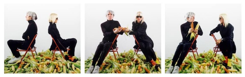Two people sit on folding chairs in a pile of corn, passing and holding ears of corn.