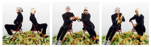 Two people sit on folding chairs among a pile of corn, handing ears of corn between them.