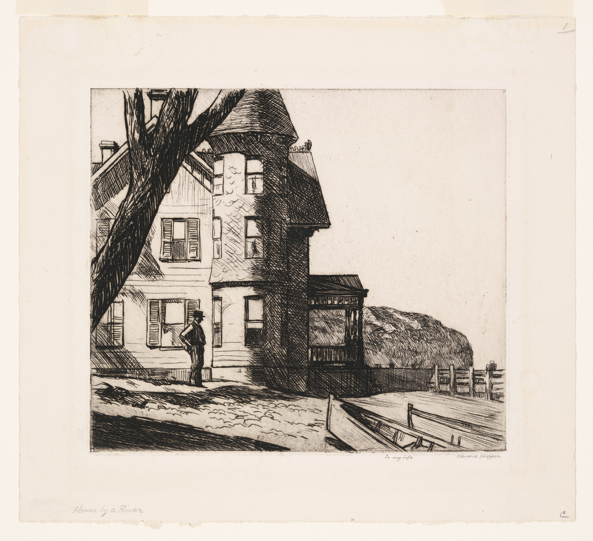 A man stands beside a large riverside house with a small boat pulled up on the shore.