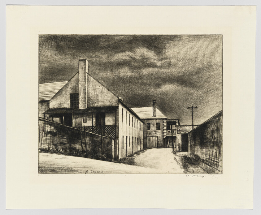 A quiet row of old industrial buildings lines an empty street under a heavy, cloudy sky.