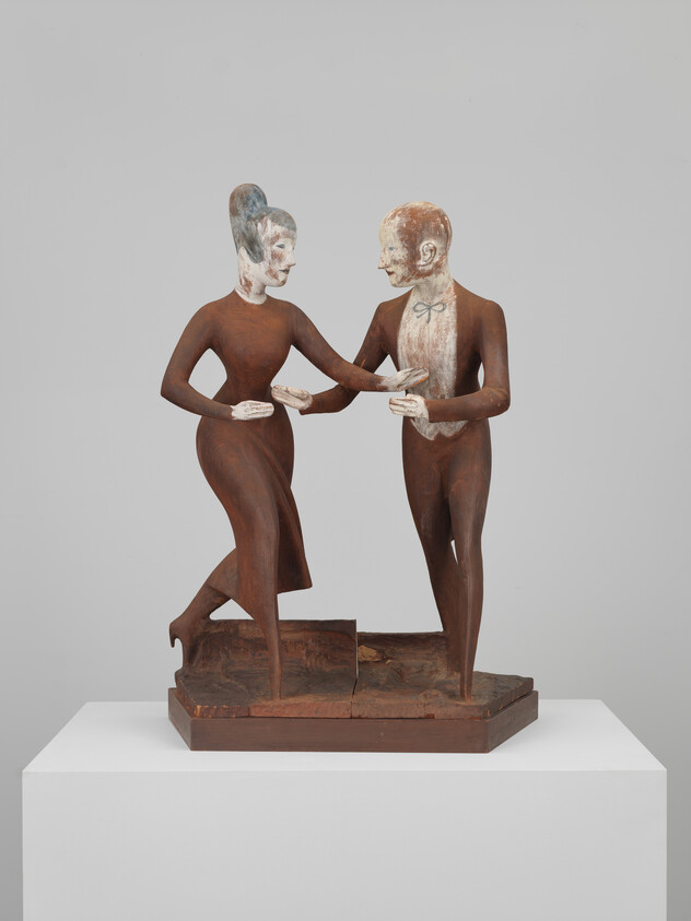 A sculpture of two stylized human figures with elongated limbs and bodies, appearing to be in a dance pose or intimate interaction. The figures have contrasting painted details, with one figure having a white face with blue details and the other with a brown face with white details. They are mounted on a wooden base, displayed against a neutral background.