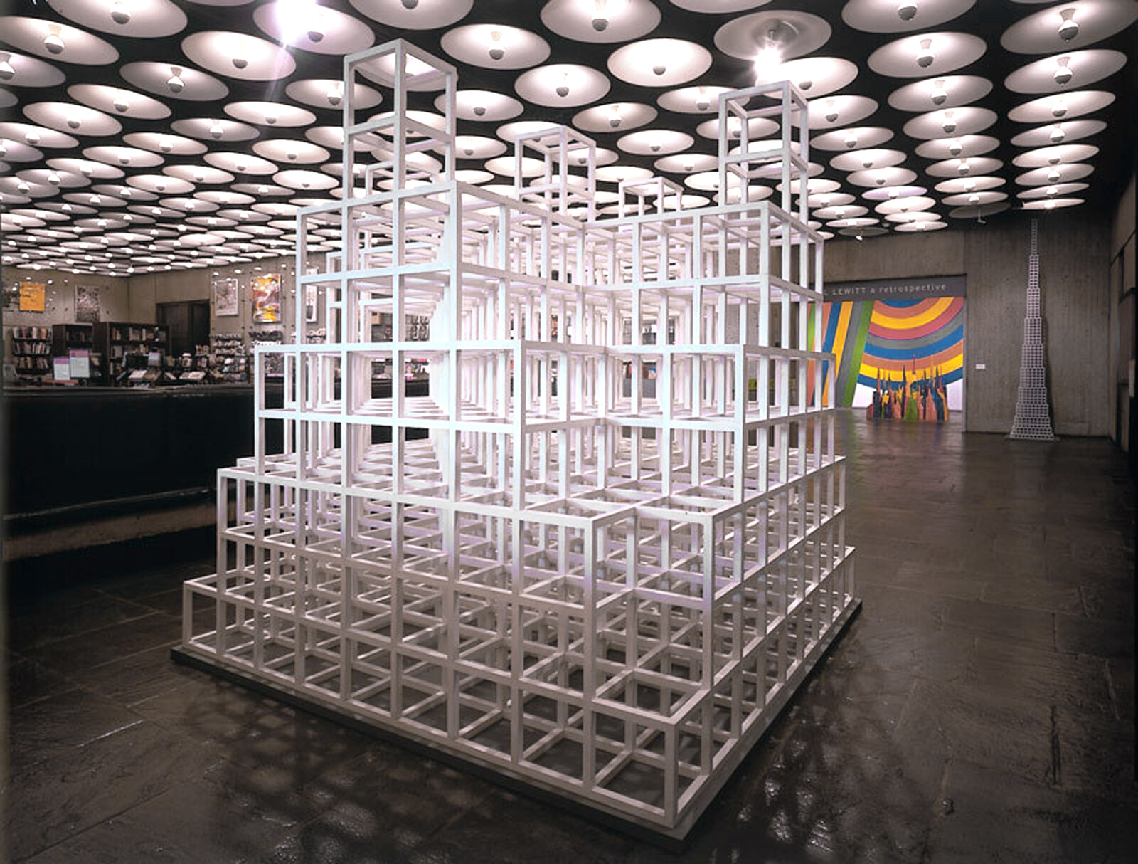 A large white cubic lattice sculpture stands in a gallery beneath a patterned round-light ceiling.