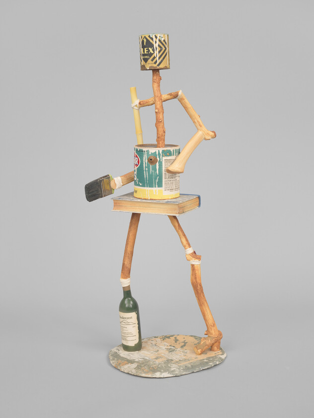 A stick-figure sculpture made from a paint can, brush, book, bottle, and wooden branches.