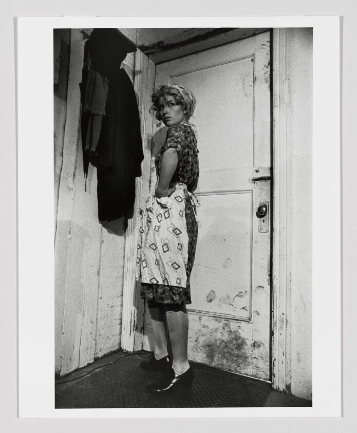 Woman in an apron stands by a worn door, looking over her shoulder.