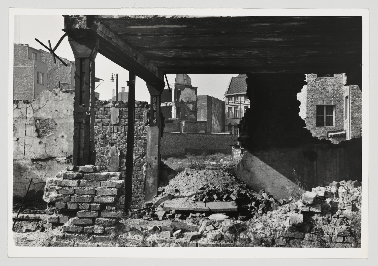 A black and white photograph showing the ruins of a building with crumbling walls and exposed beams. In the background, intact buildings can be seen, suggesting a contrast between destruction and intact urban structures. The foreground is littered with debris and broken bricks, indicating recent demolition or decay.