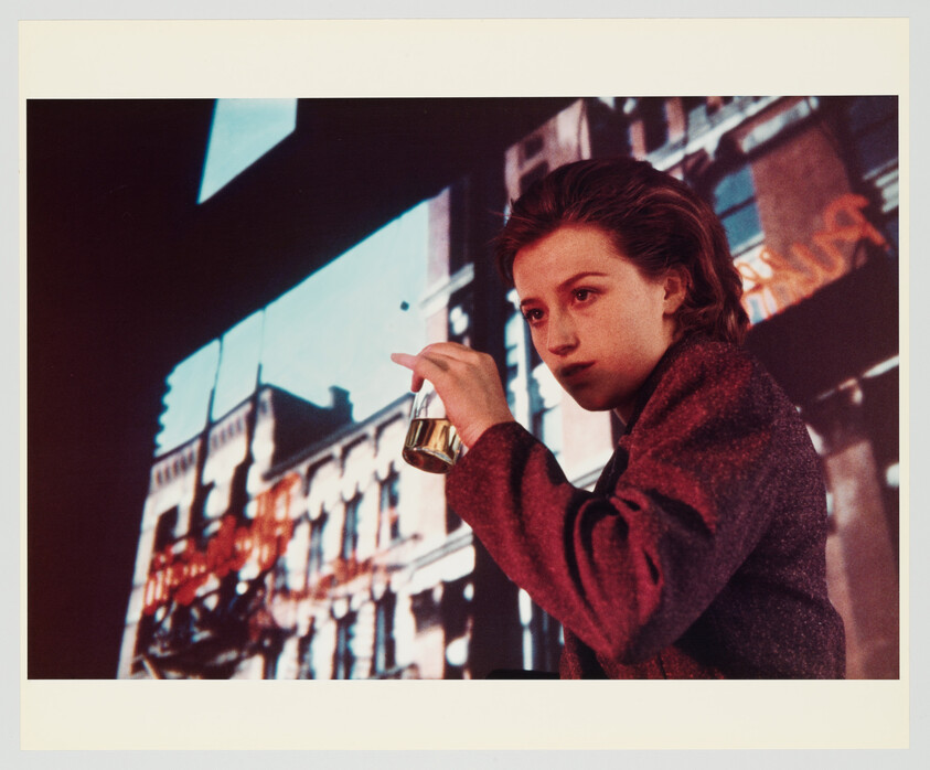 A young person holds a small glass of drink while standing before a projected city building.
