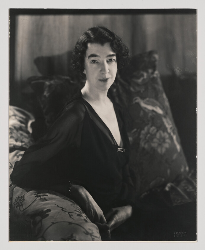 A woman in a dark dress sits on a patterned sofa, looking calmly toward the camera.