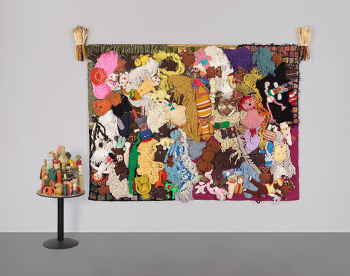 A colorful textile wall hanging covered with knitted dolls and fabric scraps beside a small pedestal sculpture.