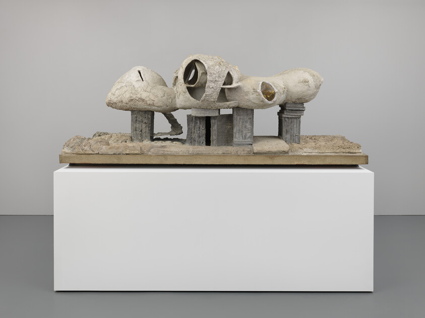 A sculpture resembling a fragmented architectural model is displayed on a white pedestal against a grey background. The sculpture features three rounded, dome-like structures with openings, supported by a series of vertical columns, all rendered in a textured, concrete-like material.