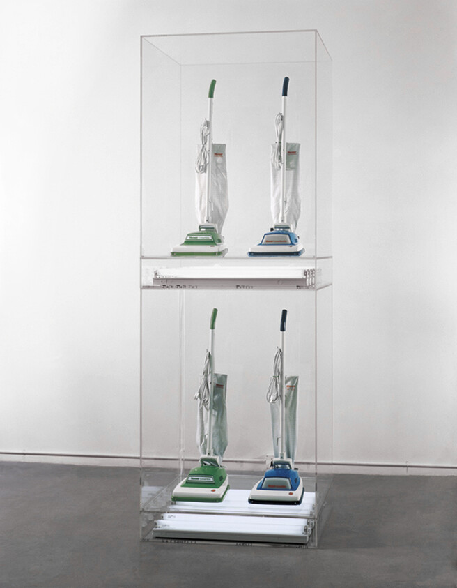 Four upright vacuum cleaners, two green and two blue, displayed on two clear shelves inside a transparent case.