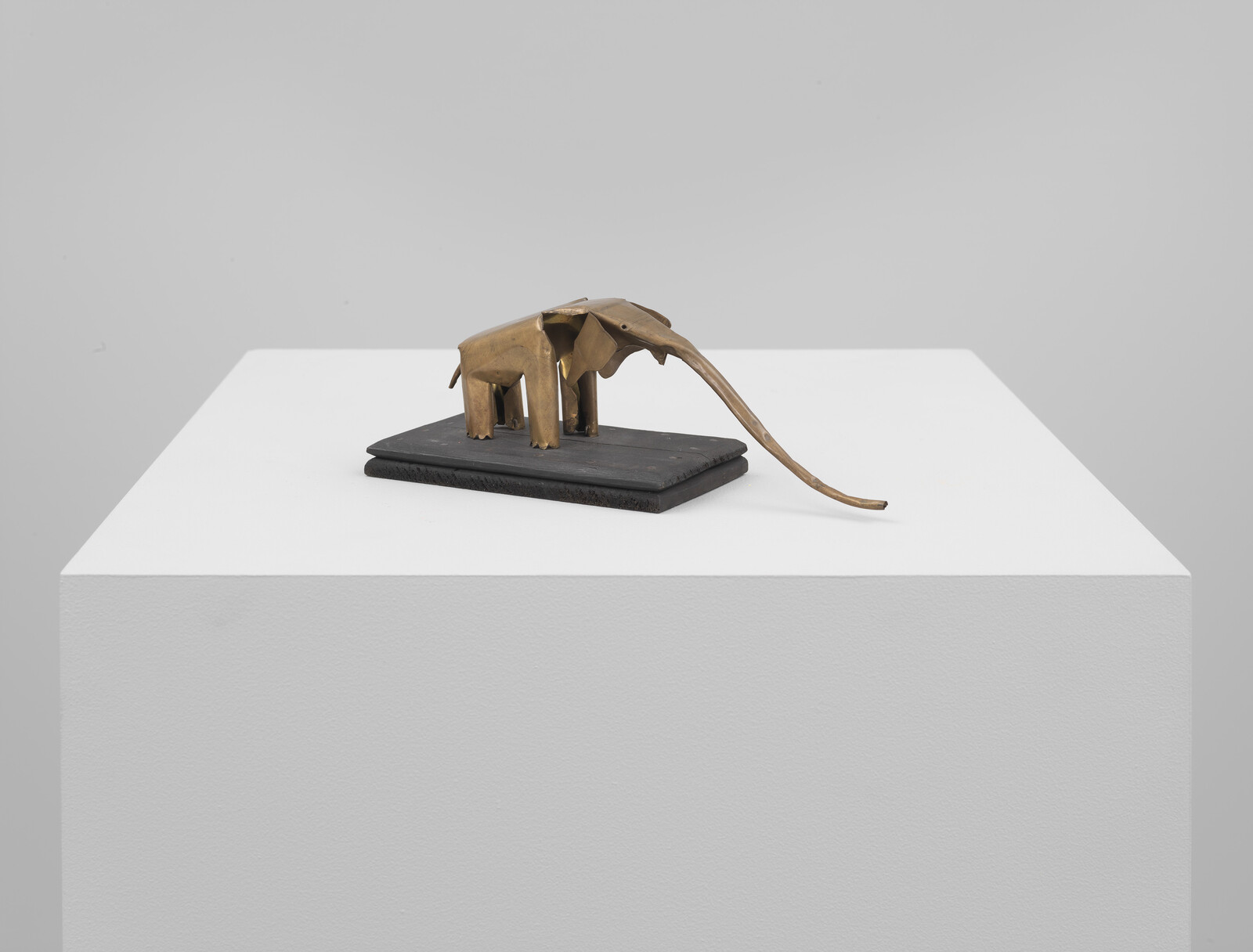 Small brass elephant sculpture with an exaggerated long curved trunk displayed on a white pedestal.