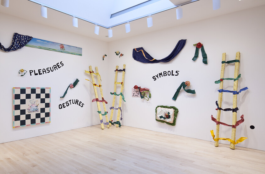 White gallery wall displaying colorful ladders and fabric ribbons around the words pleasures, gestures, and symbols.