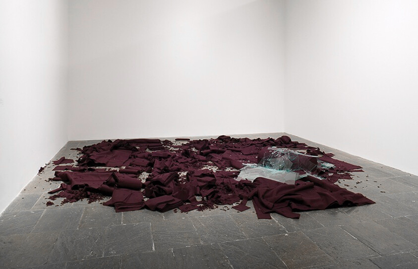 Burgundy fabric shredded and scattered on a gallery floor around a pile of broken glass.