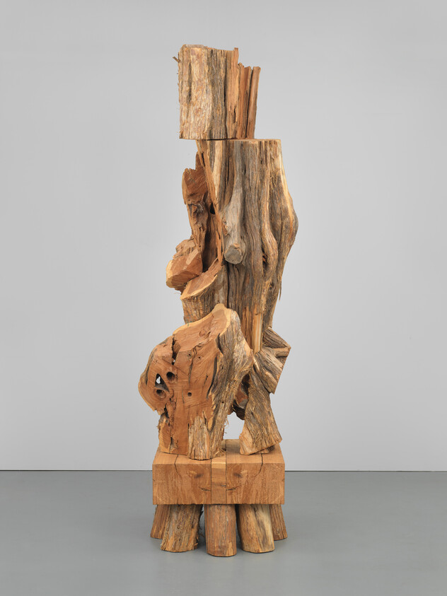 Tall abstract sculpture made from rough stacked tree trunks and blocks standing on a gallery floor.