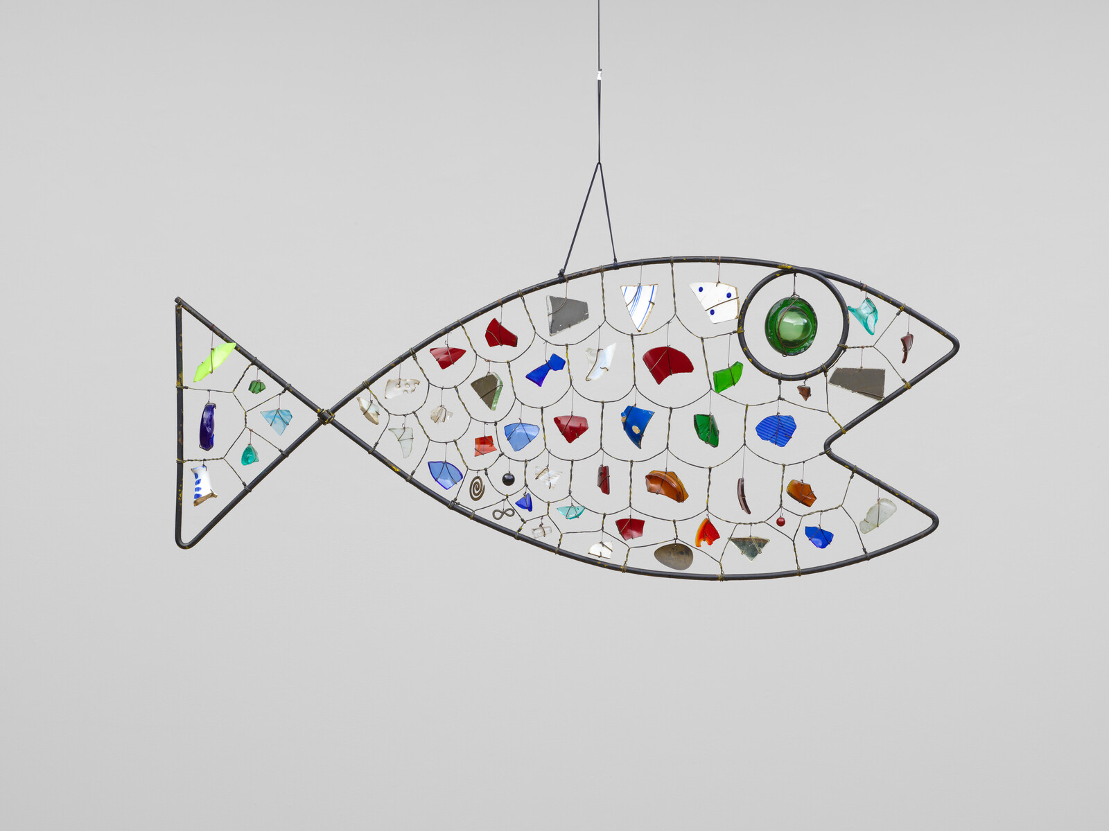 Wire fish shape filled with colorful pieces of glass and plastic, hanging against a plain light background.