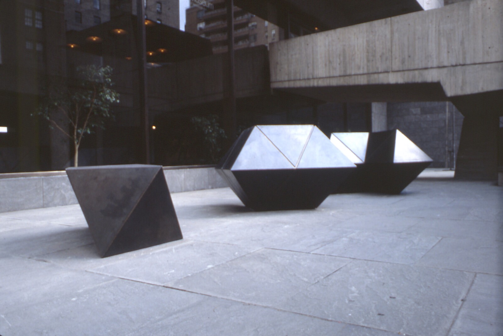 Three large dark geometric metal sculptures sit on a stone plaza under a concrete overhang.