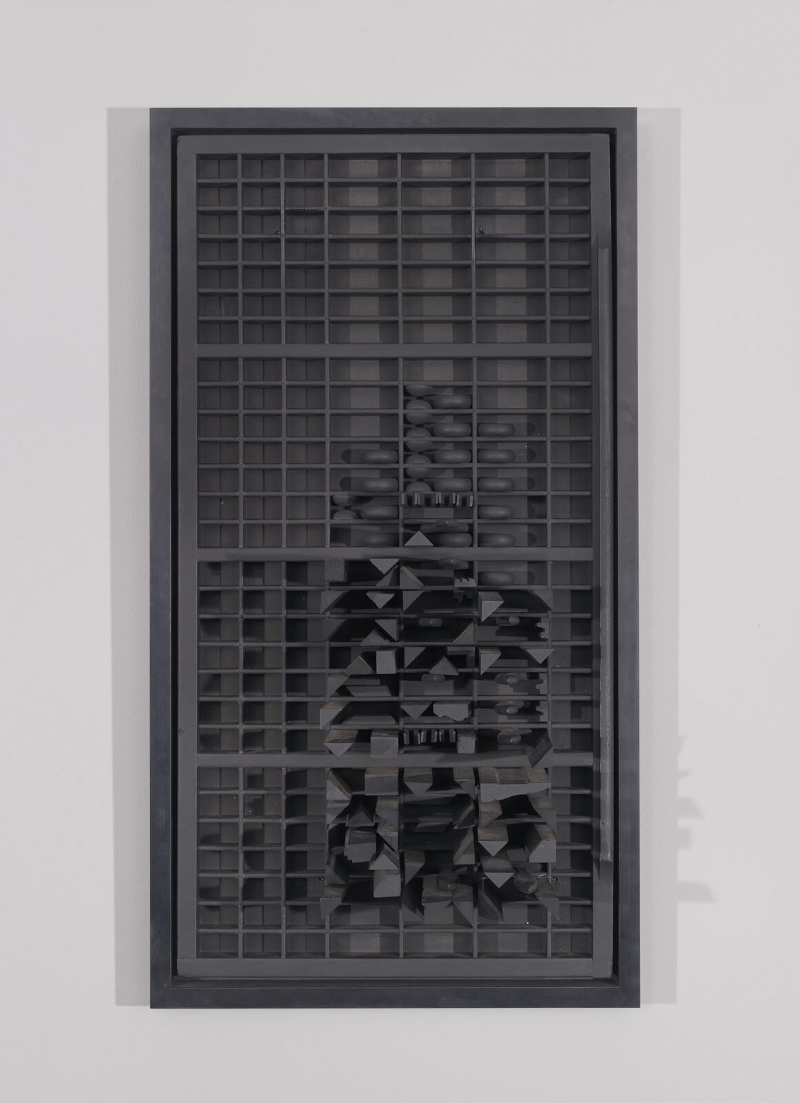 A tall black grid sculpture mounted on the wall with stacked and angular black blocks inside.