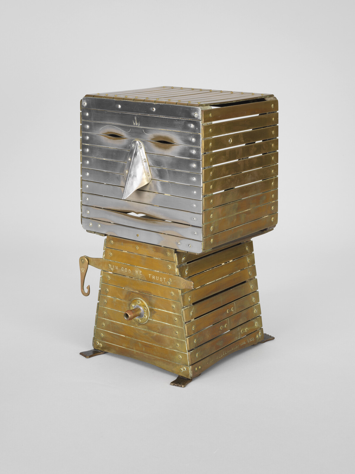Brass slatted box sculpture forms a stylized face on a pedestal inscribed with "In God We Trust."