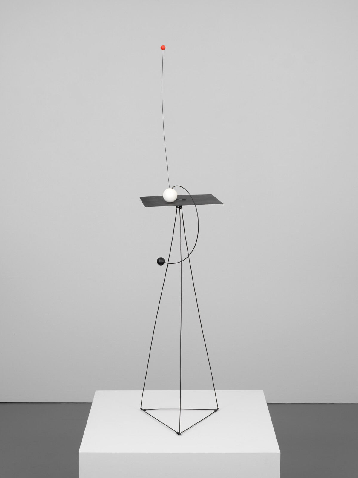 Thin black wire sculpture on a white plinth with a flat plate, white and red spheres.