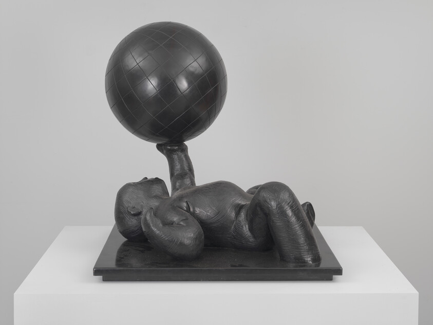 A bronze baby sculpture lies on its back holding a large patterned ball above its chest.
