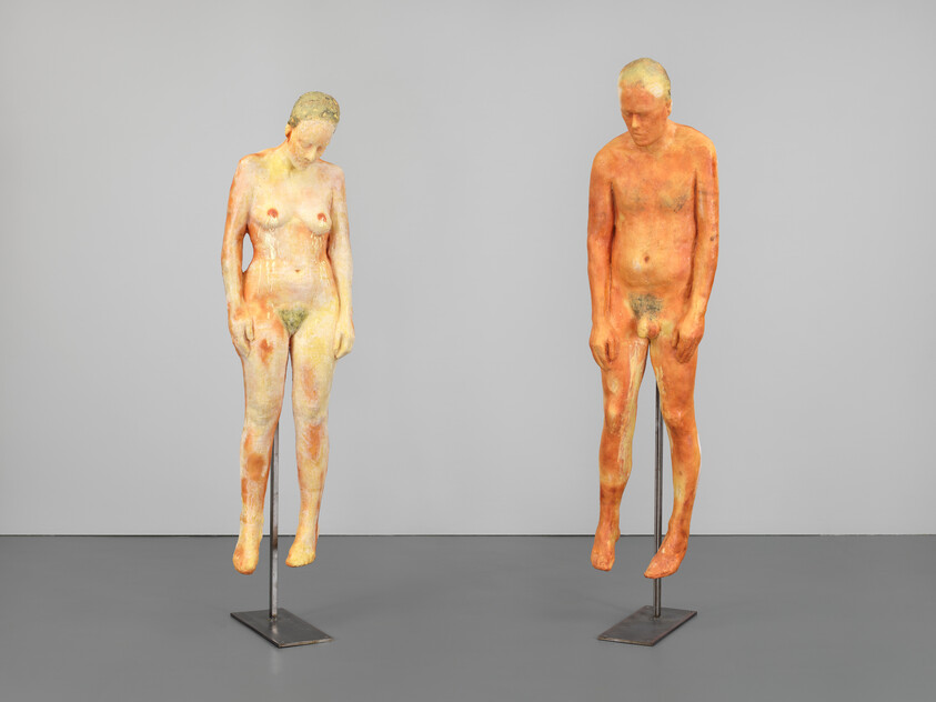 Two life-size nude sculptures of a man and a woman suspended on metal stands, heads bowed.