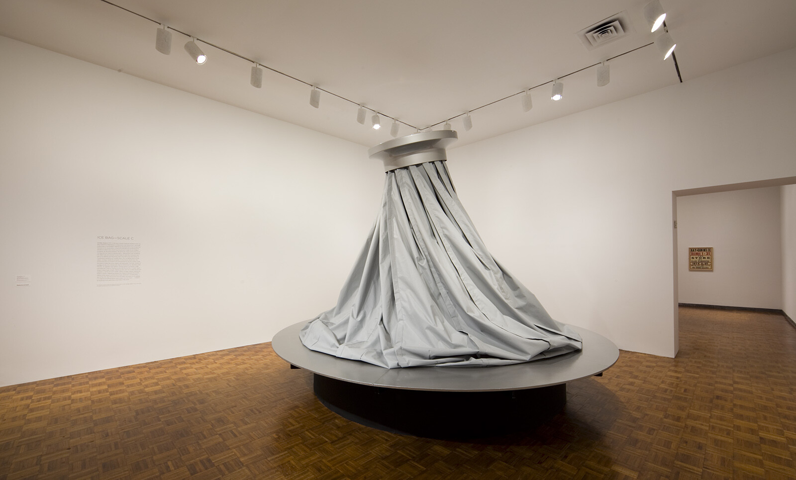 Large gray fabric drapes from a ceiling fixture onto a circular platform in an empty gallery room.