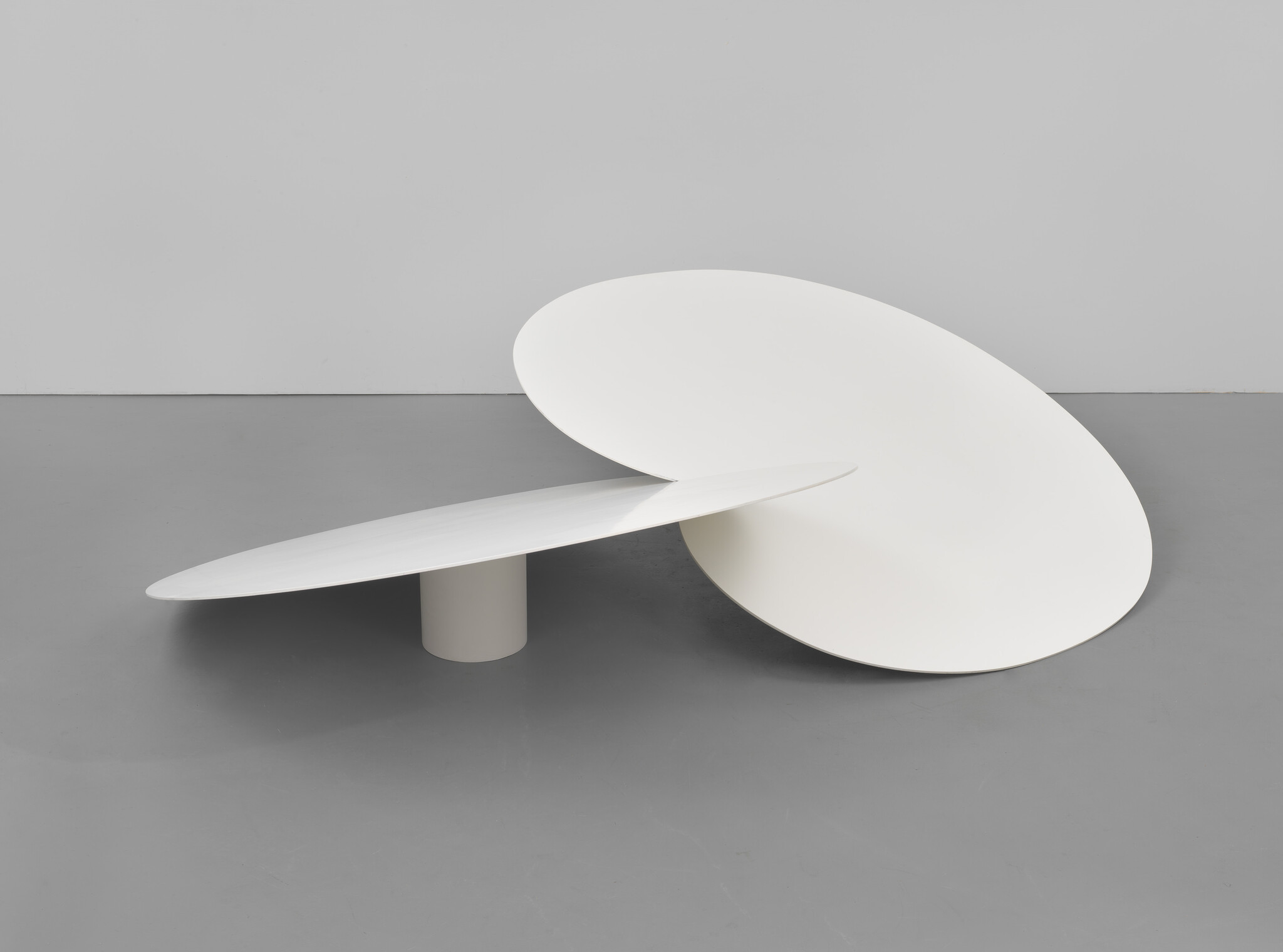 Two large white elliptical discs form a minimalist sculpture, one disc leaning against the other on a gray floor.