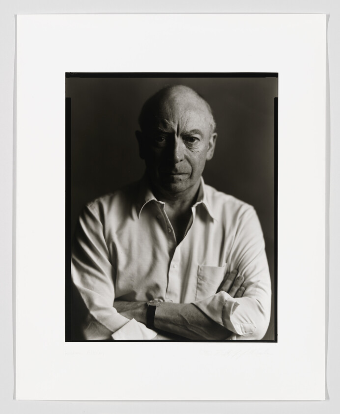 Black and white portrait of a bald man with a serious expression, wearing a white shirt with sleeves rolled up, arms crossed, and a watch on his left wrist. The photo is framed with a white border containing a signature at the bottom right.