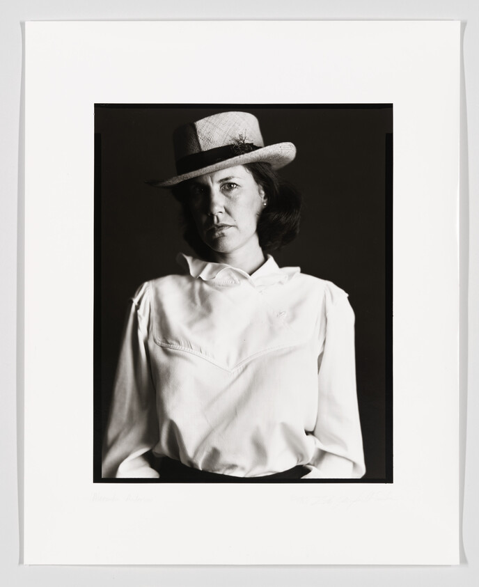 A black and white portrait of a woman wearing a wide-brimmed hat and a white blouse, with a serious expression on her face. The photograph is framed with a white border, and there is a signature at the bottom.