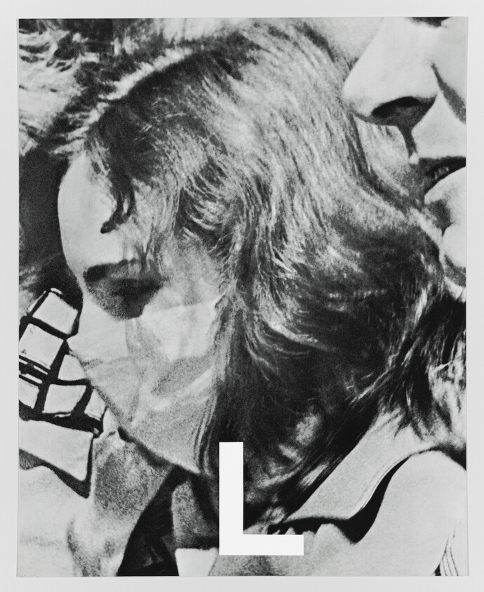 Close-up of two people pressed together, one wearing a face covering, large letter L at bottom.