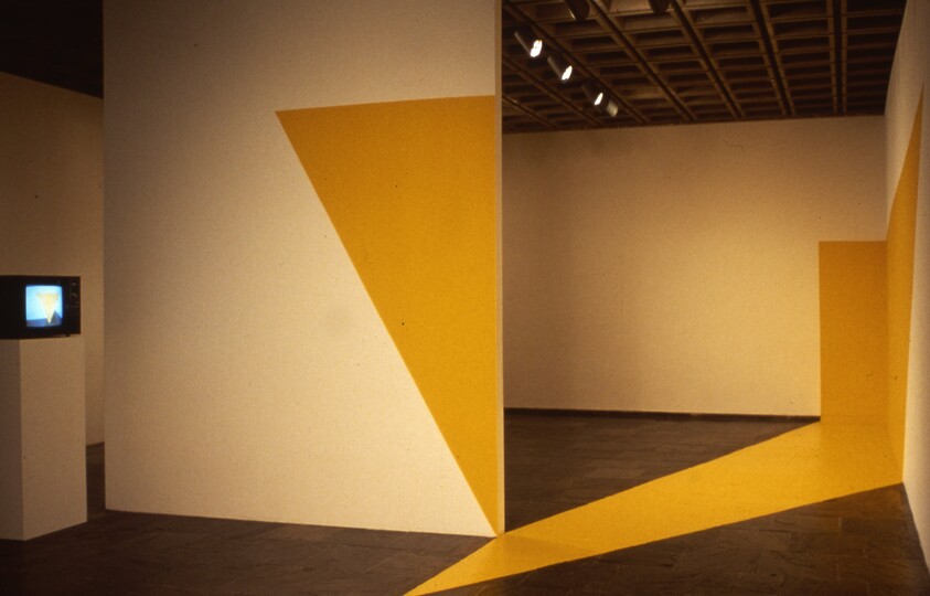 Yellow triangular shapes painted across gallery walls and floor with a small TV on a pedestal.