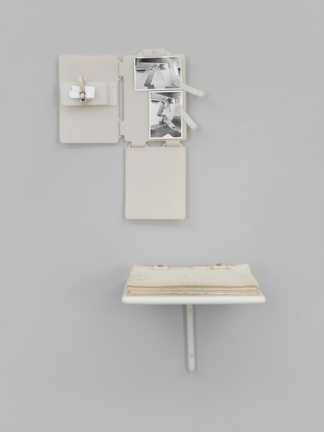 Minimal wall installation with clipped black-and-white photos above a small shelf holding stacked papers.