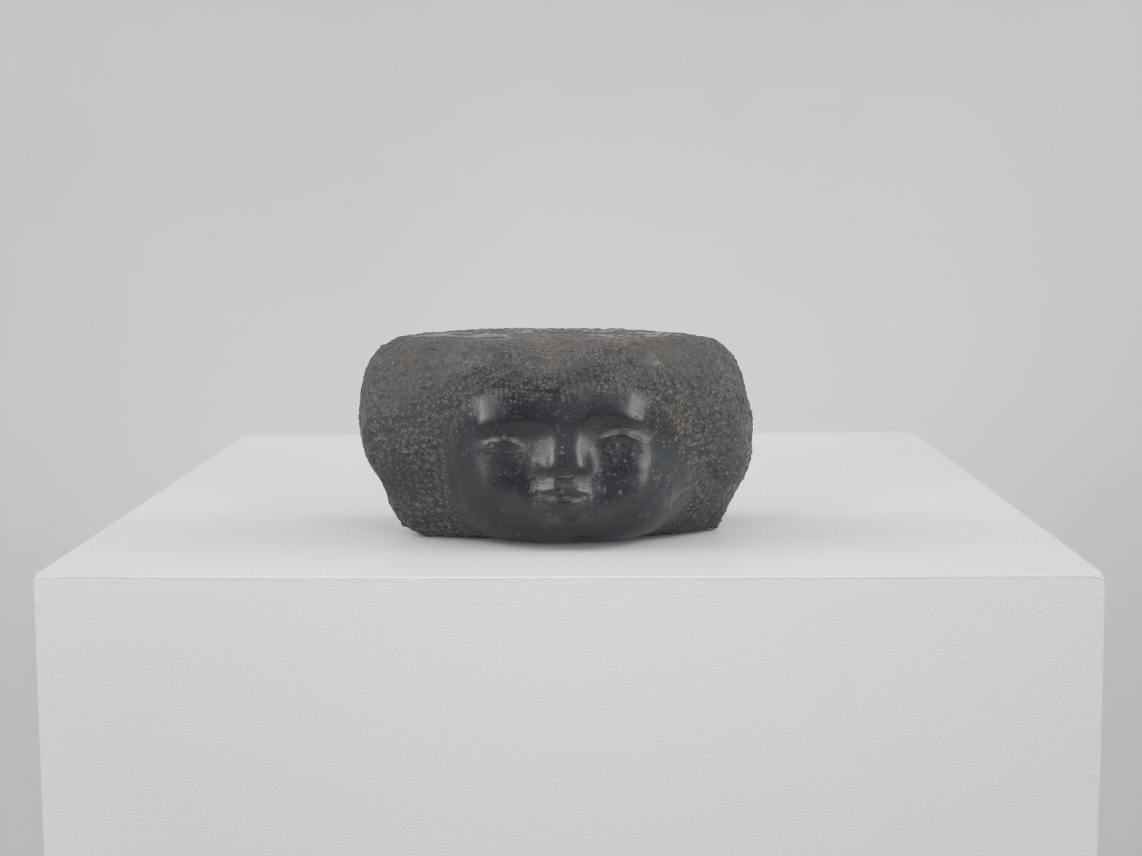 A stone sculpture with a minimalist human face rests on a white pedestal against a light grey background. The sculpture appears weathered and has a rough texture, with the facial features smoothly carved into the stone.