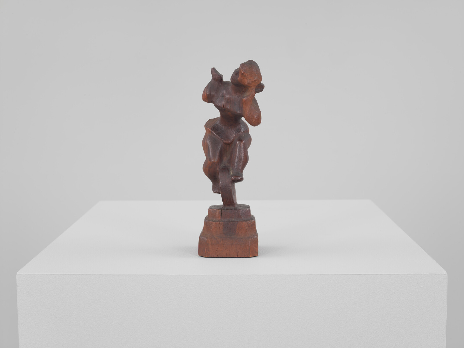 A small carved wooden figurine with abstract human features stands on a white pedestal.